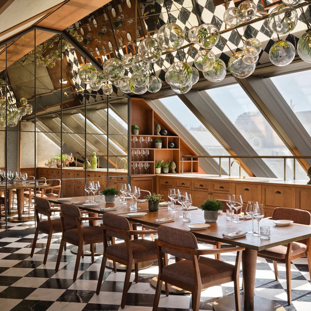 A dining table under hanging glass lights and  large slanted windows.